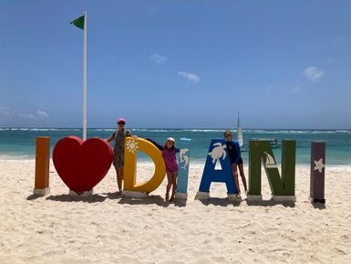 Drei Personen stehen am Strand vor bunten Buchstaben, die 'I ♥ Diani' bilden, während das Meer im Hintergrund glitzert.