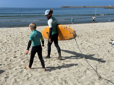 Ein Surflehrer und ein Junge gehen am Strand entlang, während der Lehrer ein großes, orangefarbenes Surfbrett trägt.