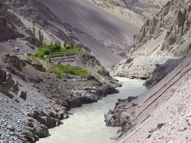 Der Indusfluss schlängelt sich durch eine schroffe, bergige Landschaft, umgeben von steilen Felsen und grünen Terrassenfeldern.