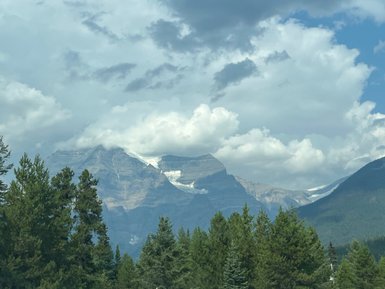 Majestätische Berge erheben sich hinter dichten, grünen Wäldern unter einem bewölkten Himmel mit sanften Lichtstrahlen.