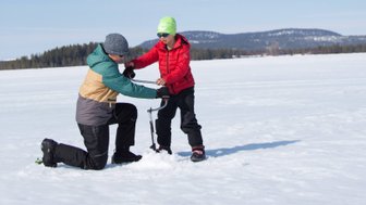 Ein Erwachsener und ein Kind stehen auf einer schneebedeckten Fläche, während sie gemeinsam ein Loch ins Eis bohren.