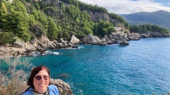 Eine Person mit Brille lächelt vor einer malerischen Küstenlandschaft mit klarem Wasser und grünen Hügeln.