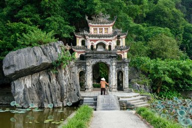 Ein Mann mit einem Stock steht vor einem alten, bunten Tempel, umgeben von üppigem Grün und einem ruhigen Teich.