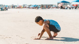 Ein Junge mit lockigem Haar sitzt am Strand und gräbt mit seinen Händen im Sand, während die Sonne scheint.