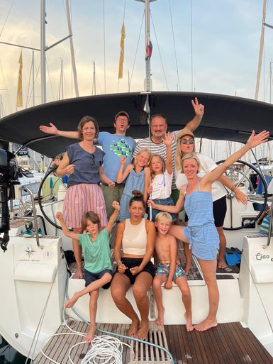Eine fröhliche Gruppe von Erwachsenen und Kindern posiert auf einer Segelyacht, umgeben von Masten und einem schönen Himmel.