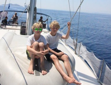 Zwei Jungen mit blonden Haaren sitzen auf dem Bug eines Segelbootes, umgeben von glitzerndem Wasser und klarem Himmel.
