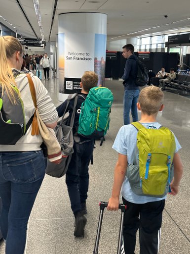Eine Familie mit Rucksäcken betritt den Flughafen, während ein Willkommensschild für San Francisco sichtbar ist.