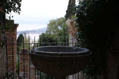 Ein steinerner Brunnen steht im Vordergrund, umgeben von einer Eisenzaun und üppigem Grün, mit einem Blick auf die Küste im Hintergrund.