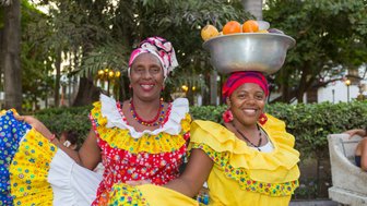 Zwei fröhliche Frauen in bunten traditionellen Kleidern posieren in einem Park, eine trägt eine Schüssel mit Früchten auf dem Kopf.