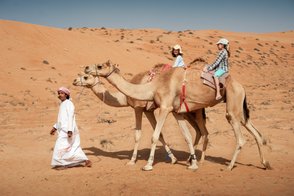 Kinder reiten gemeinsam mit einem Einheimischen auf Kamelen – Oman Reise mit Kindern