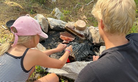 Ein Mädchen in einem gestreiften Shirt grillt über einem Lagerfeuer, während ein Junge daneben zuschaut und sich konzentriert.