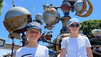 Zwei Kinder stehen vor einem bunten, futuristischen Karussell in Disneyland, umgeben von strahlend blauem Himmel.