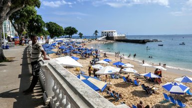 Ein Mann lehnt an einem Geländer und beobachtet den Strand, wo sich viele Menschen unter blauen Sonnenschirmen versammeln.