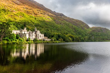 Ein majestätisches Schloss spiegelt sich im ruhigen Wasser eines Sees, umgeben von üppigem, grünem Wald und sanften Hügeln.