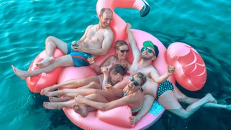 Fünf Freunde entspannen auf einem großen, pinken Flamingo-Schwimmring im klaren Wasser, lachen und genießen Getränke.