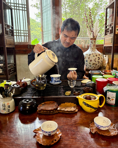 Ein Mann gießt dampfenden Tee aus einer weißen Kanne in eine kleine Tasse, umgeben von traditionellem Teezubehör.