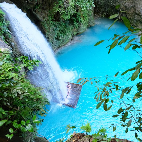Ein Wasserfall stürzt in ein leuchtend blaues Wasserbecken, umgeben von üppigem Grün und tropischen Pflanzen.