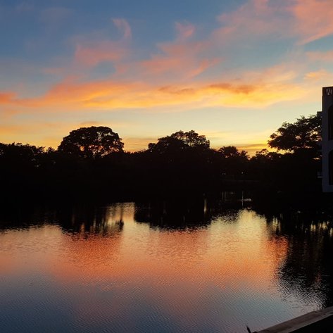 Ein atemberaubender Sonnenuntergang spiegelt sich im ruhigen Wasser eines Sees, umgeben von üppigem Grün und Bäumen.