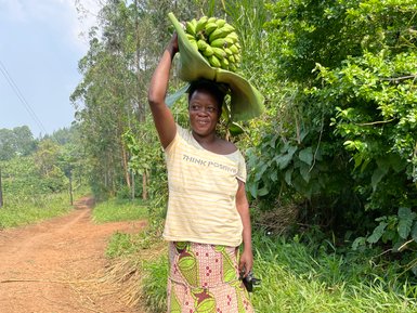 Eine Frau trägt eine große Ladung Bananen auf ihrem Kopf, während sie auf einem staubigen Weg in der Natur steht.