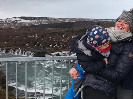 Eine lachende Frau hält ein kleines Kind in den Armen, während sie vor einem Wasserfall steht, umgeben von einer malerischen Landschaft.