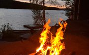 Ein loderndes Lagerfeuer brennt in einer Feuerschale, umgeben von Bäumen und einem ruhigen See im Hintergrund.