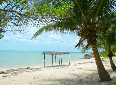 Ein ruhiger Strand mit feinem Sand, umgeben von Palmen und einem kleinen, schattigen Unterstand am Wasser.