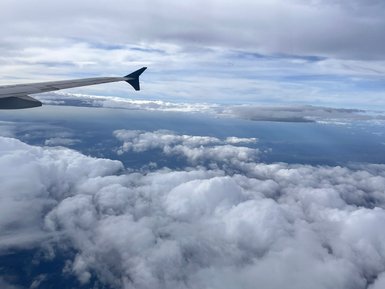 Der Blick aus dem Flugzeug zeigt die Wolkenlandschaft unterhalb, mit einem Flugzeugflügel im Vordergrund.