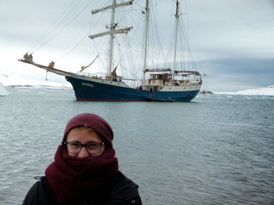 Ein Mann mit einer warmen Mütze und einem dicken Schal steht am Wasser, während ein großes Segelschiff im Hintergrund liegt.
