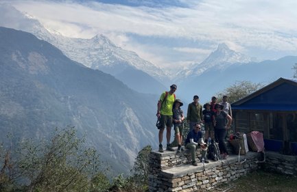 Eine Gruppe von Wanderern posiert auf einer Steinmauer mit schneebedeckten Bergen im Hintergrund und strahlendem Himmel.