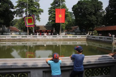Zwei Kinder stehen am Ufer eines ruhigen Teichs und beobachten die Umgebung, während bunte Fahnen im Hintergrund wehen.
