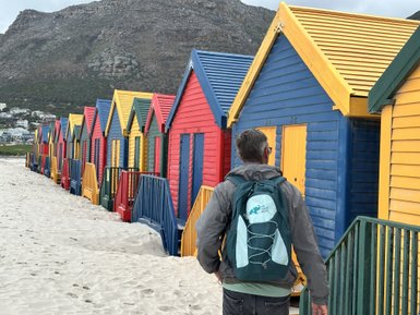 Ein Mann mit einem Rucksack geht an bunten Strandhütten entlang, die in verschiedenen Farben gestrichen sind.