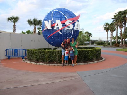 Eine Familie posiert fröhlich vor dem NASA-Symbol, umgeben von Palmen und einem gepflegten Garten.