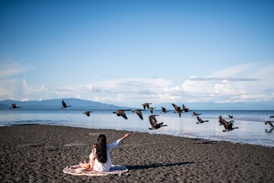 Eine Frau sitzt auf einem Teppich am Strand und beobachtet eine Gruppe von Gänsen, die über das Wasser fliegen.