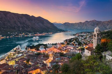 Ein atemberaubender Blick auf die Bucht von Kotor, umgeben von majestätischen Bergen und beleuchteten Städten bei Sonnenuntergang.