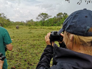 Eine Frau mit einer Kamera beobachtet ein Nashorn in der Wildnis, während ein Mann im Hintergrund steht.
