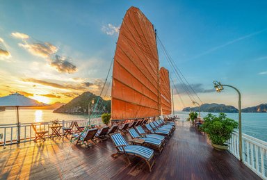 Die Terrasse eines Schiffes in der Ha Long Bucht mit orangefarbenen Segeln und Liegestühlen, während die Sonne untergeht.