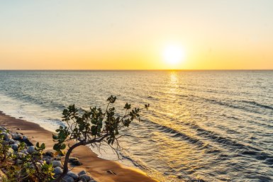 Ein ruhiger Strand mit sanften Wellen, während die Sonne am Horizont untergeht und den Himmel in warmen Farben erleuchtet.