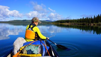 Ein Junge paddelt in einem Kanu auf einem ruhigen See, umgeben von grünen Bäumen und einem strahlend blauen Himmel.