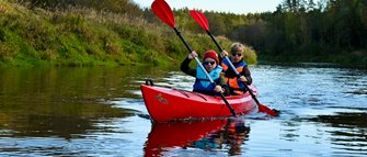 Zwei Kinder paddeln in einem roten Kajak auf einem ruhigen Fluss, umgeben von grünen Ufern und Bäumen im Hintergrund.