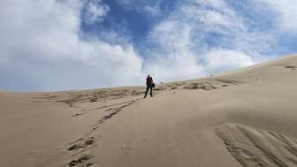 Ein Mensch erklimmt eine hohe Sanddüne in der Mongolei, umgeben von einem weiten, wolkigen Himmel.