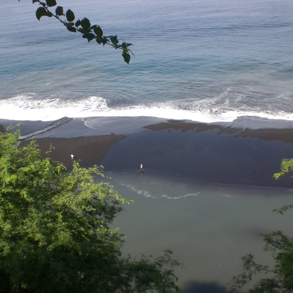 Ein Blick auf den ruhigen Ozean, wo sanfte Wellen an den schwarzen Sandstrand rollen, umgeben von üppigem Grün.
