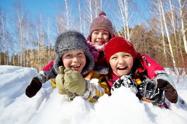 Drei fröhliche Kinder lachen im Schnee, während sie sich in warmen Winterkleidern umarmen und die Sonne genießen.