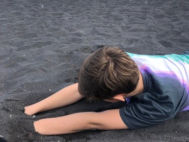 Ein Junge liegt auf schwarzem Sand und gräbt mit seinen Händen, während das Meer sanft im Hintergrund plätschert.
