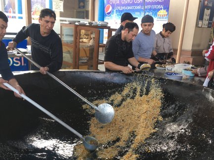 Mehrere Männer rühren in einem großen Wok mit Reis und Gemüse, während sie in einem belebten Markt in Taschkent arbeiten.