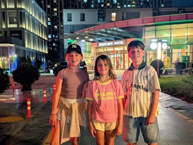 Drei Kinder stehen fröhlich vor dem Mercure Hotel in Beijing bei Nacht, umgeben von bunten Lichtern.