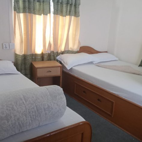 Ein einfaches Zimmer mit zwei Einzelbetten, einem Nachttisch und Vorhängen, die sanft das Licht filtern.