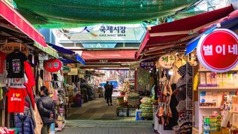 Ein belebter Markt mit bunten Ständen, die eine Vielzahl von Waren und Souvenirs anbieten, unter einem grünen Sonnenschutz.
