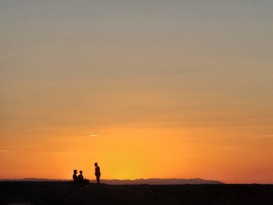 Drei Personen sitzen und stehen silhouettenhaft vor einem atemberaubenden Sonnenuntergang über den Sanddünen in der Mongolei.