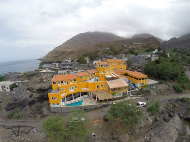 Blick auf ein leuchtend orangefarbenes Gebäude mit Pool, umgeben von sanften Hügeln und dem Meer im Hintergrund.