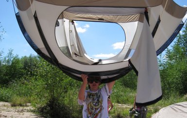 Ein Kind hält ein Zelt über dem Kopf, während es auf einem sandigen Ufer steht, umgeben von Bäumen und Zelten.